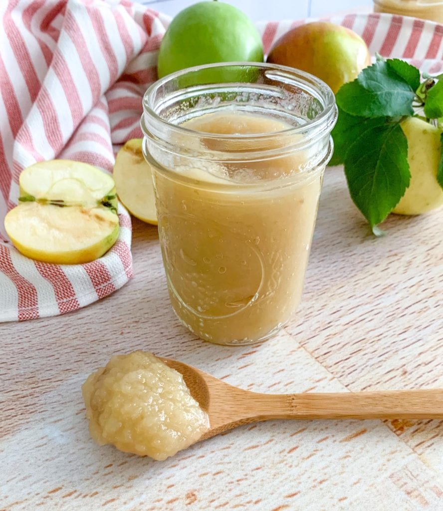 A jar of applesauce. A wooden spoon and apples in the background.