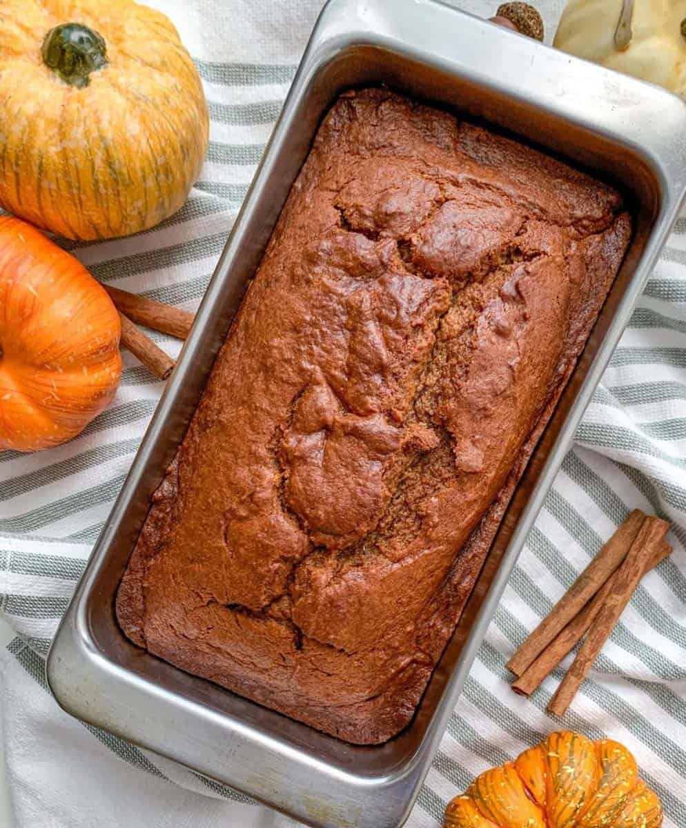 Baked pumpkin bread in a loaf plan. Decorative pumpkins and cinnamon sticks surrounding the bread.