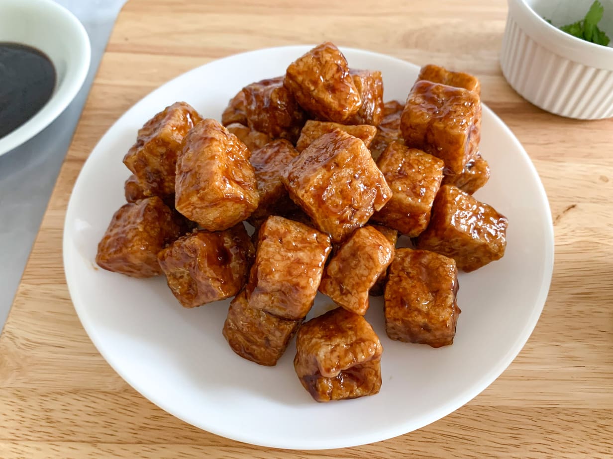 A plate of fried tofu cubes, tossed in hoisin sauce. A dish of cilantro and hoisin sauce alongside the plate.