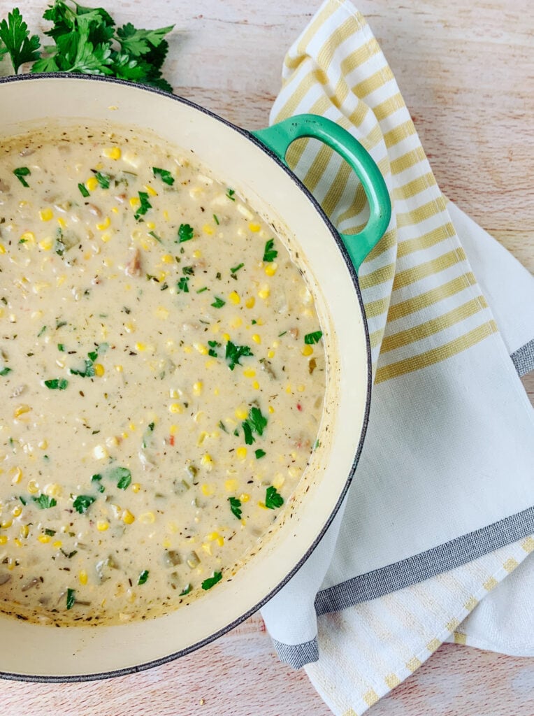 A green pot of chowder, topped with corn and herbs, A yellow striped towel around it.