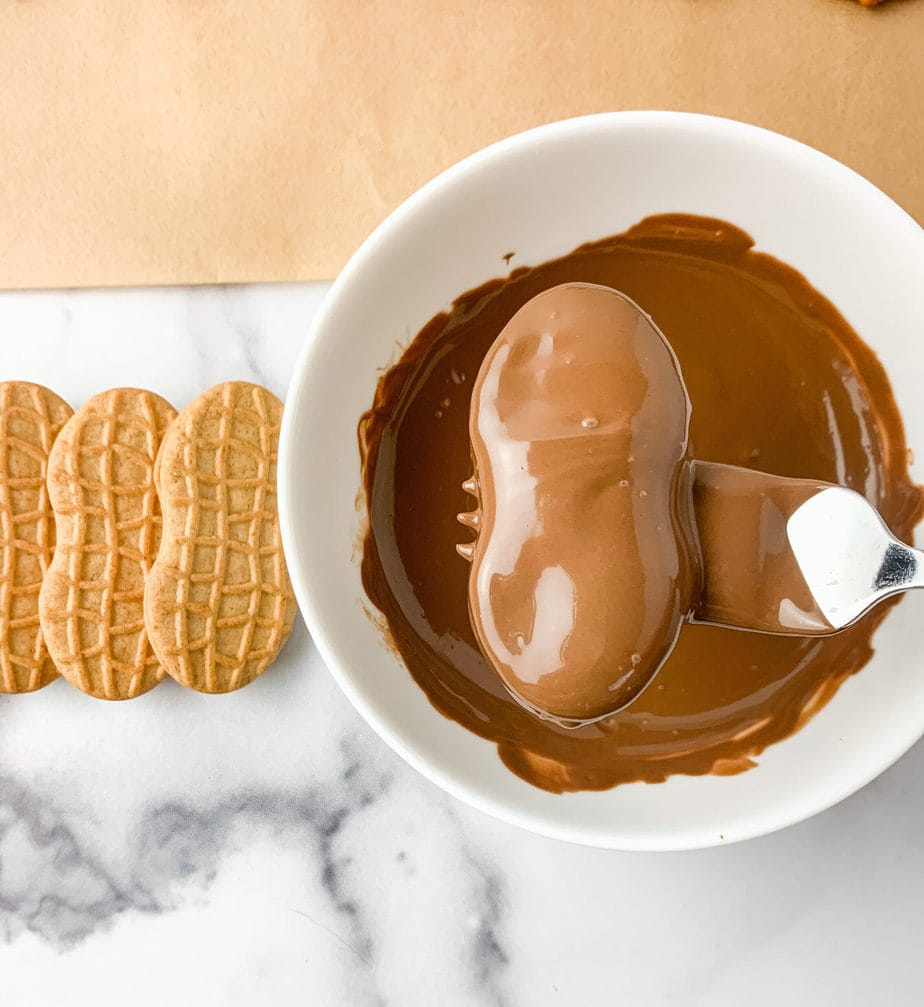 A Nutter Butter cookie dipped in chocolate being lifted by a fork. Plain Nutter Butter cookies sitting along side the chocolate.