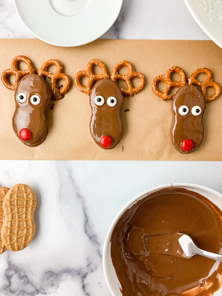 Three finished chocolate dipped Nutter Butters, decorated with two pretzel antlers, two candy eyes, and each with a red M&M nose. Setting on brown parchment paper, surrounded by white plates and a dish of melted chocolate.