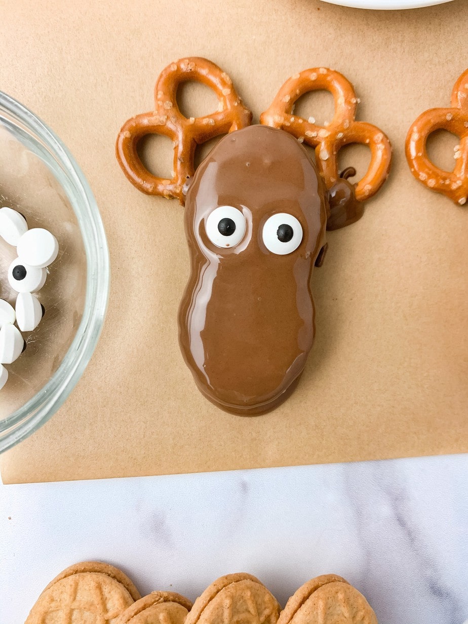 A chocolate dipped Nutter Butter Reindeer sitting on two pretzel antlers on brown parchment paper, two candy eyes added to make a face.