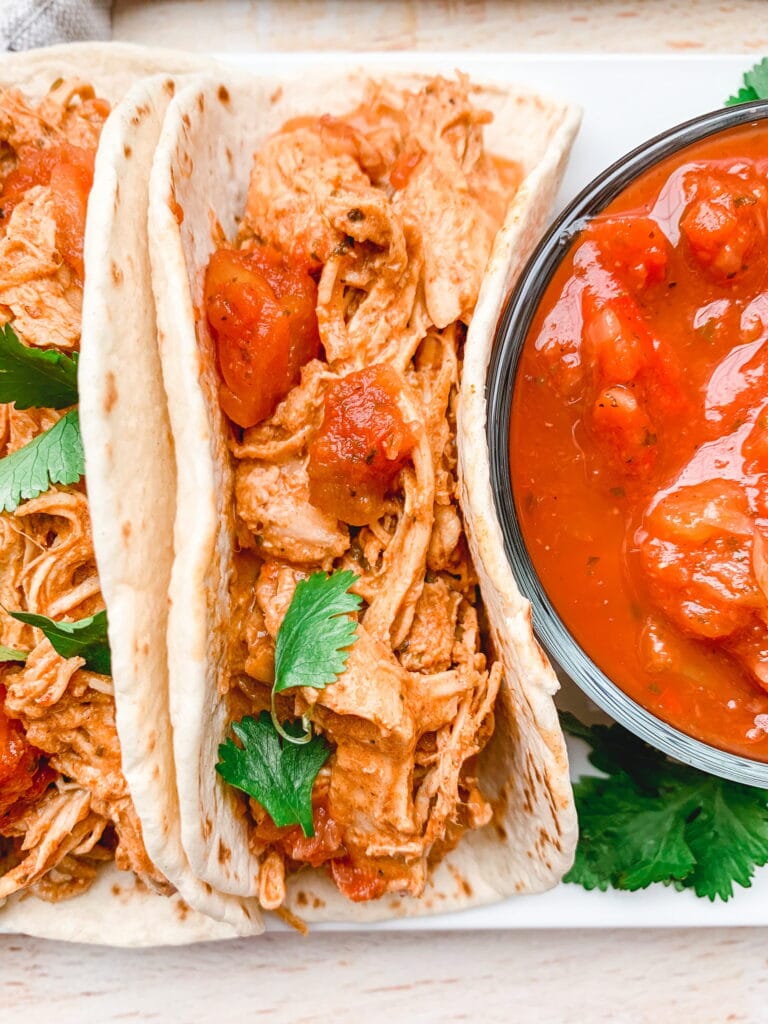 A close up of a taco with shredded chicken , tomatoes and green herbs. Salsa to the side.