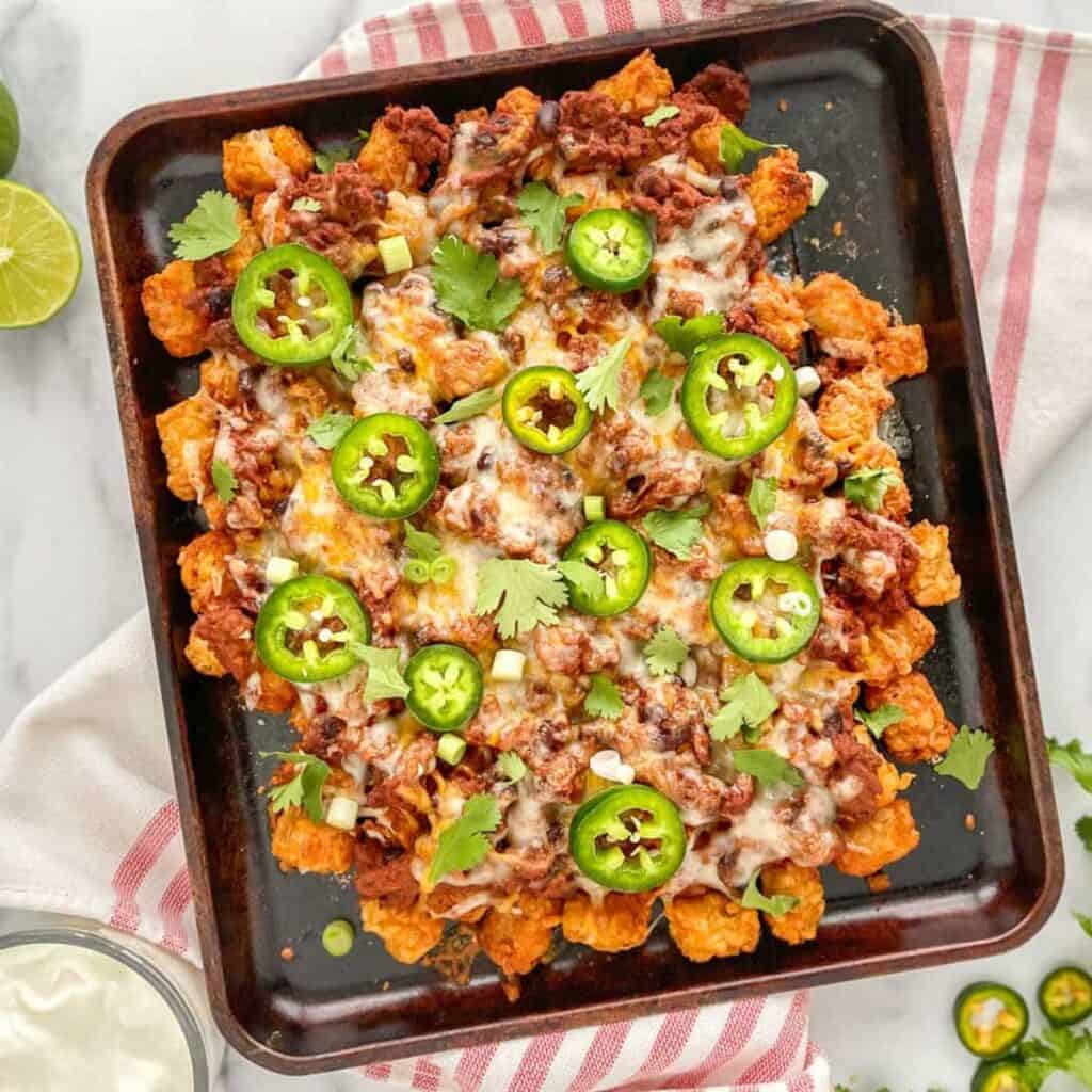 A pan of tater tots topped with chili, cheese and jalapenos. A red striped napkin, lime , and sour cream in the back