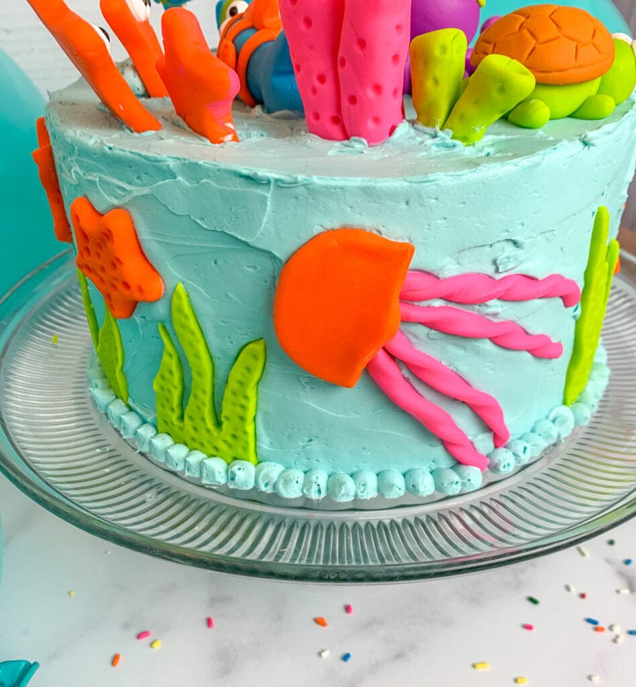 A fondant pink and orange jelly fish on the side of a light blue frosted cake with green seaweed and orange starfish made of fondant.