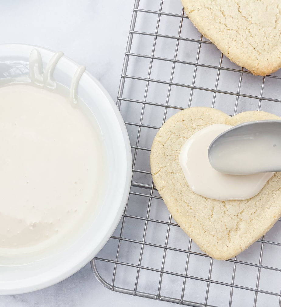 Icing a heart shaped cookie on a wire rack with white icing using a spoon. A bowl of icing is next to the cookie.