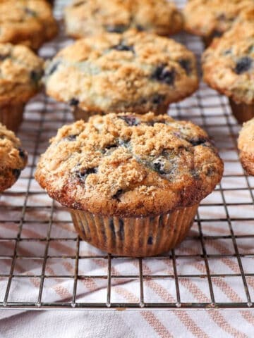 A photo of blueberry muffins on a cooling rack. A striped towel is under the rack.