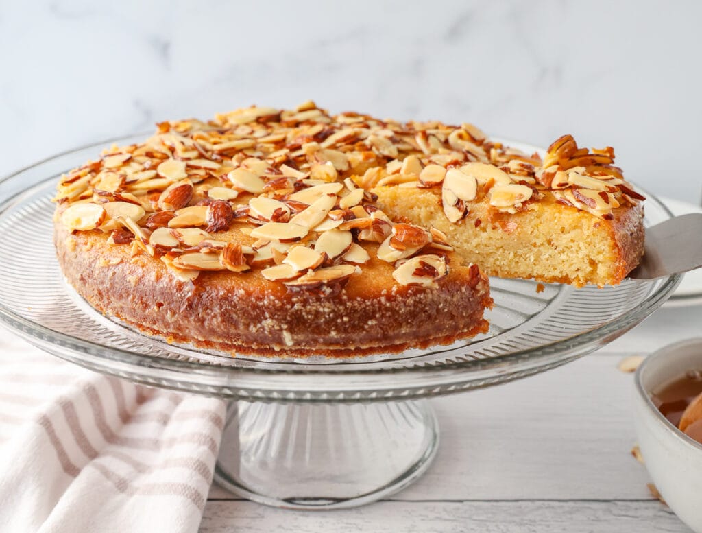 An almond cake on a cake stand with a slice being removed. 