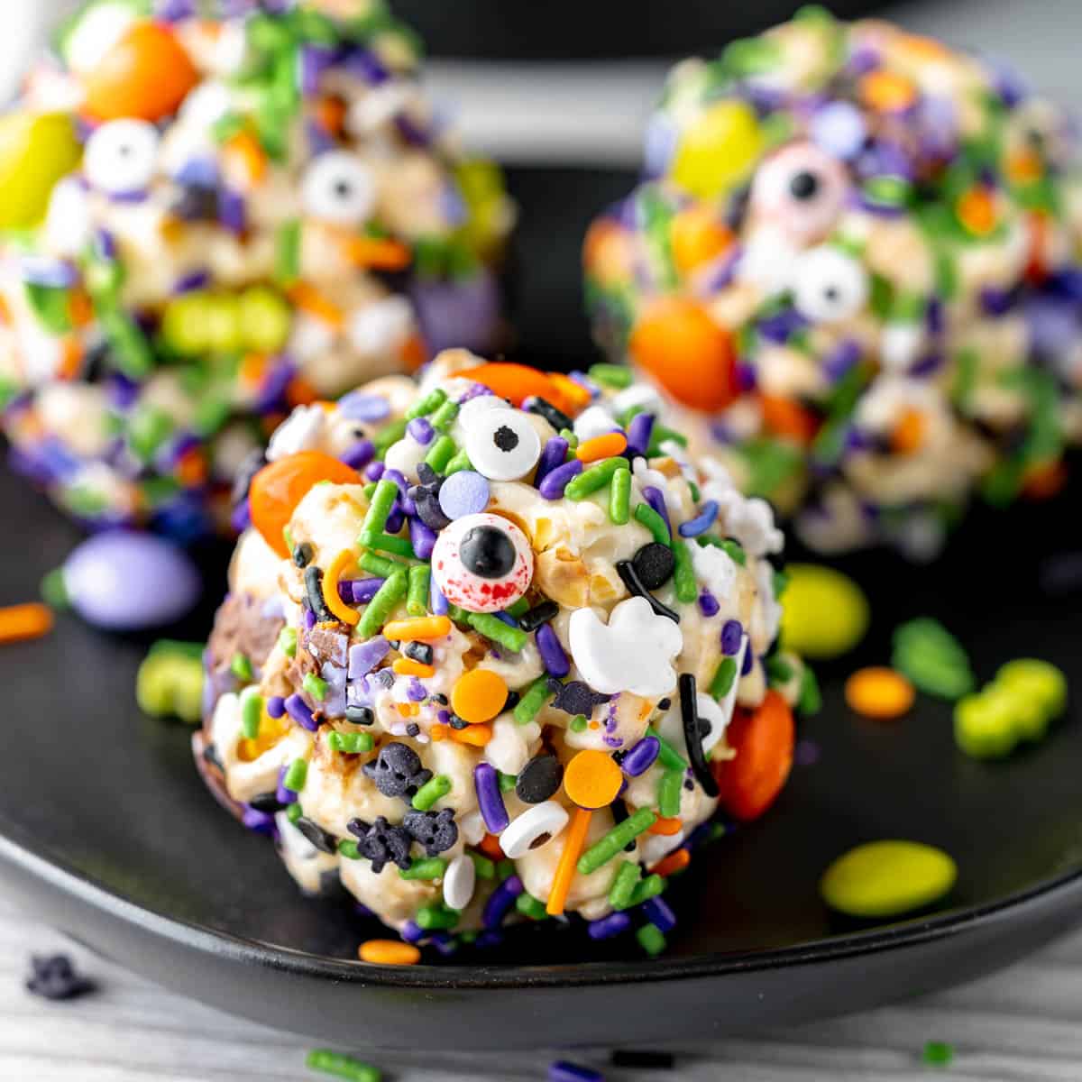 Three popcorn balls on a black plate coated in Ghoul's Mix M&Ms and Halloween Sprinkles.