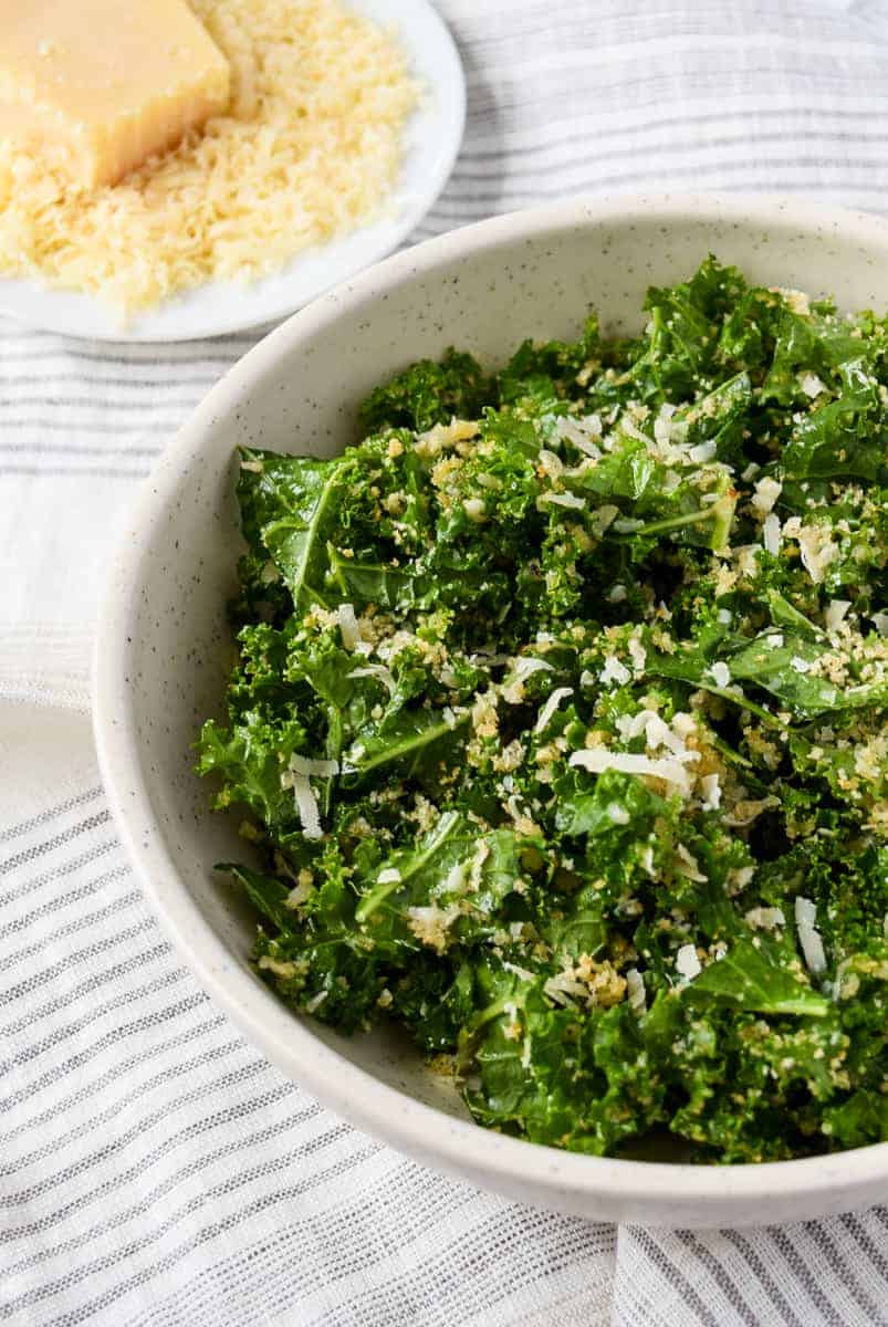 Green leafy kale in a white bowl topped with shredded parmesan cheese and toasted breadcrumbs. Parmesan cheese in a bowl sits in the background.