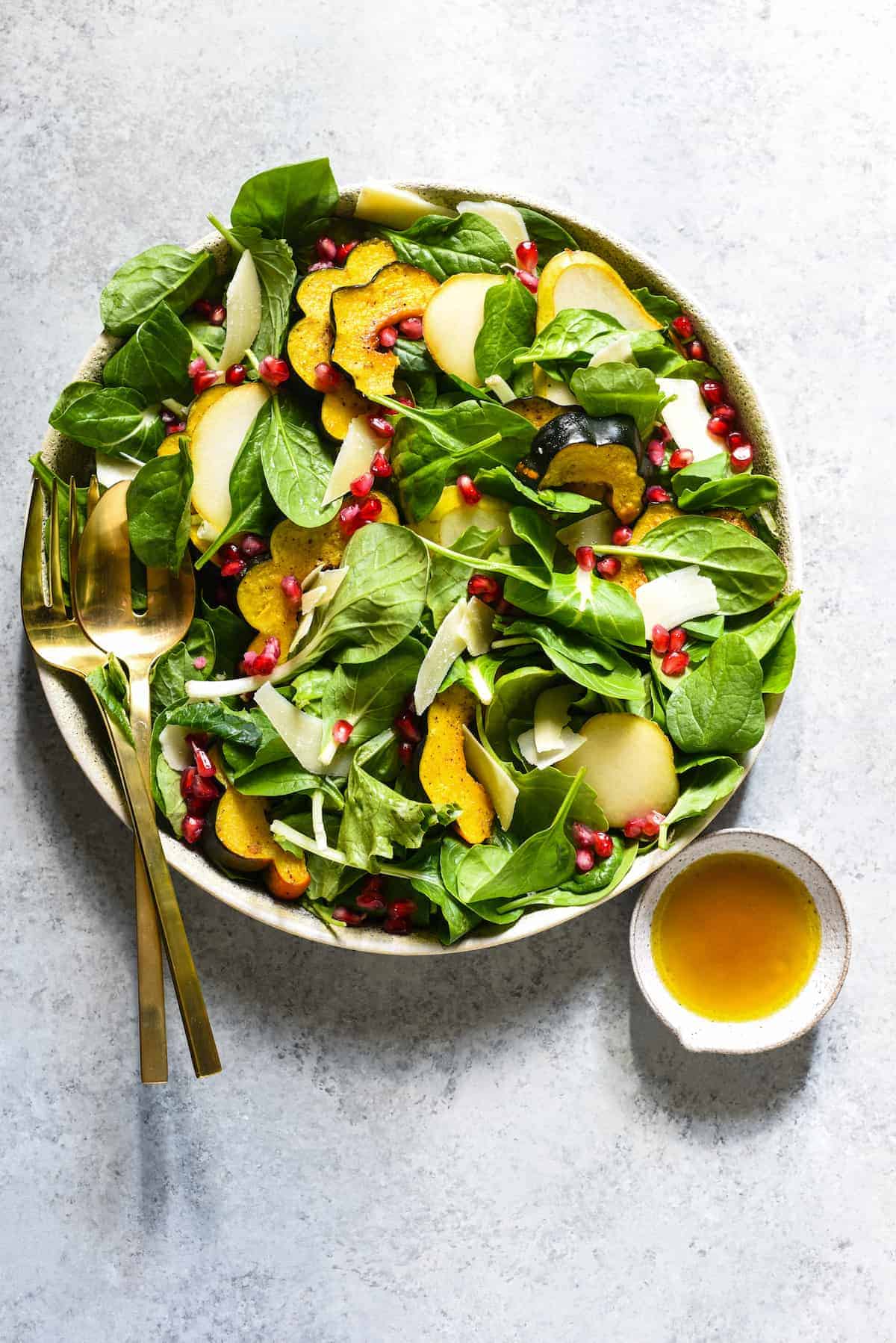 A bowl of greens in a stone bowl, topped with sliced acorn squash and pomegranate seeds. A dish of dressing and gold utensils on the side.