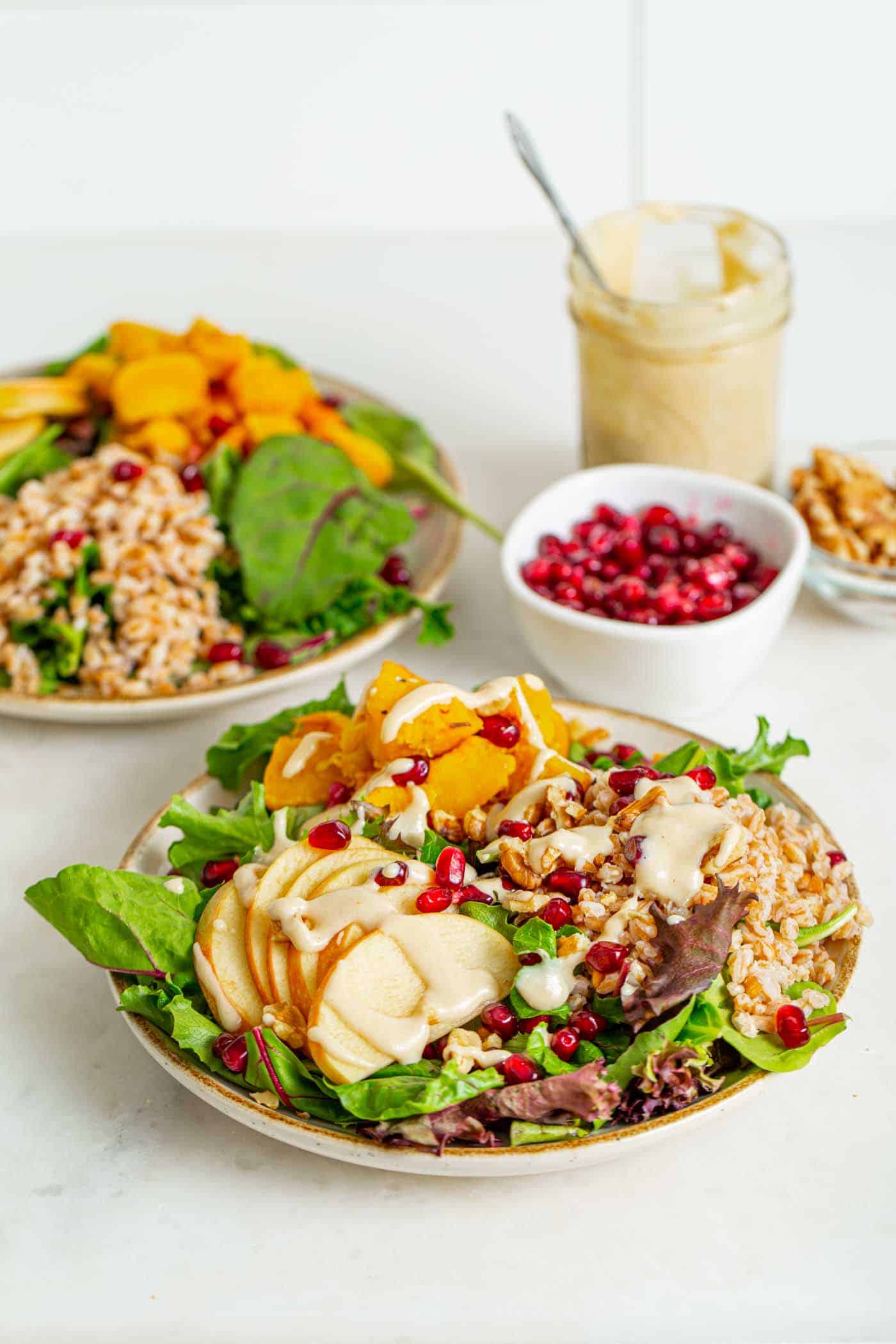 One up close shot and one far back shot of  bowls of salad, with leafy greens, sliced apples, squash, farro, topped with pomegranate seeds and creamy dressing dishes of pomegranate seeds and nuts sit in the background with a jar of extra dressing.