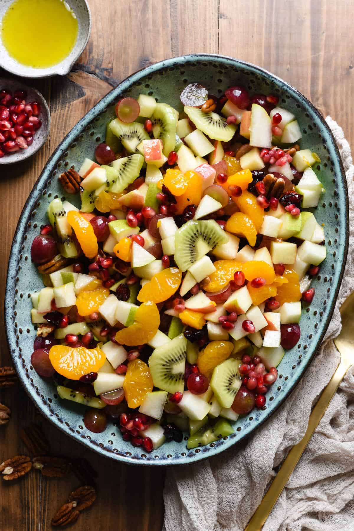 A mixture of colorful fruit is served on a blue platter. Extra dressing and pomegranate seeds are show top left, with a serving utensil sits bottom right. Extra pecans are sprinkled around the platter.