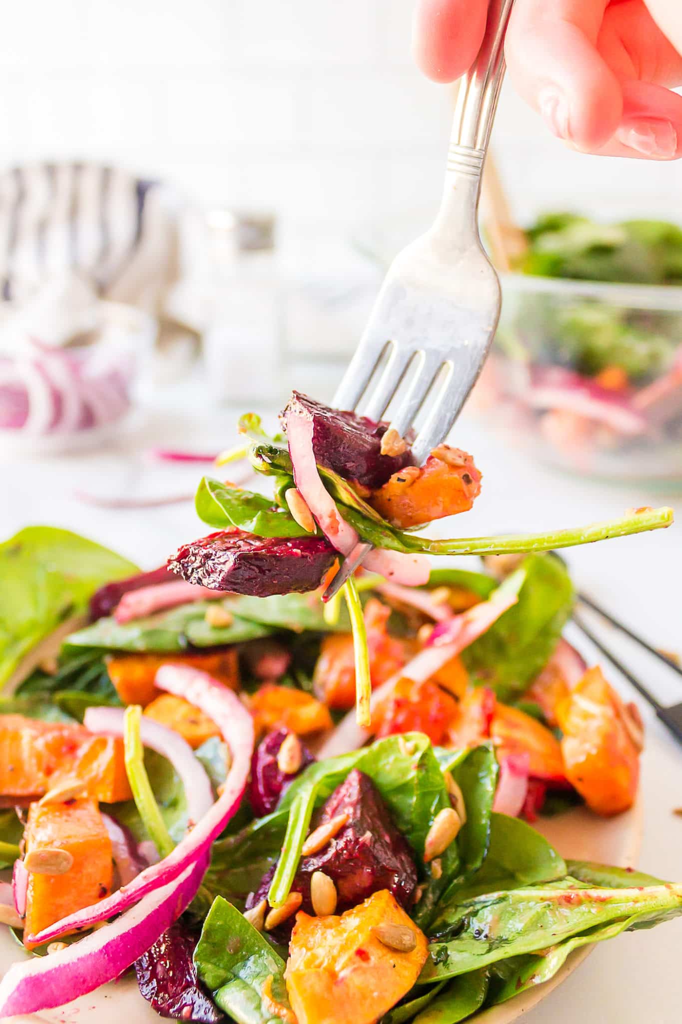 A hand with a forkful of beets, sweet potatoes,  greens, a slice of red onion and sunflower seeds, held over a plate of the salad.  A bowl of salad and the extra ingredients are in the background with a salt shaker.