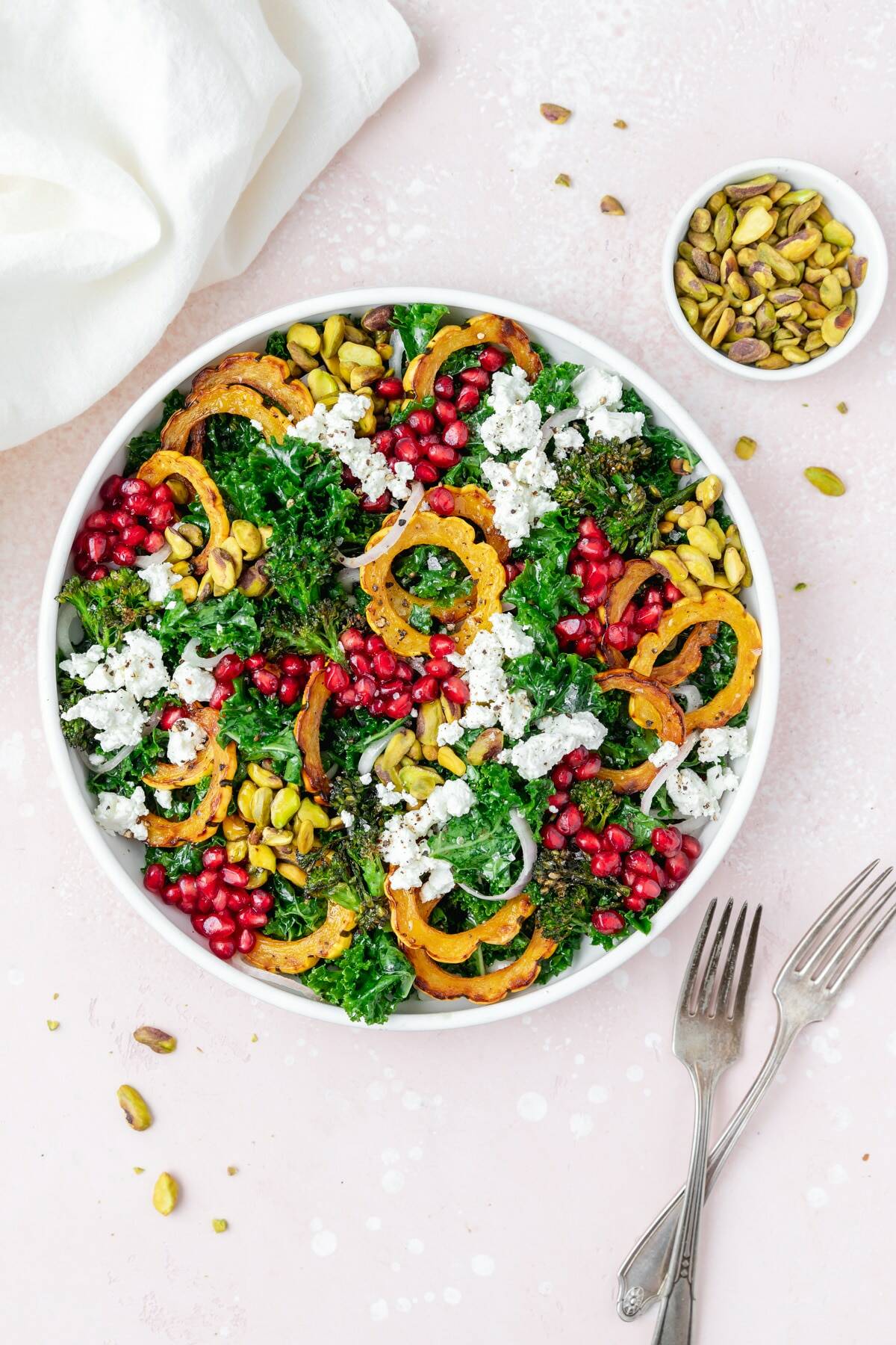 A white bowl filled with leafy green kale, slices of orange squash, pistachios, pomegranate seeds and chunks of goat cheese. A bowl of pistachios sits above the salad with a set of forks and a white napkin on each side of the bowl.