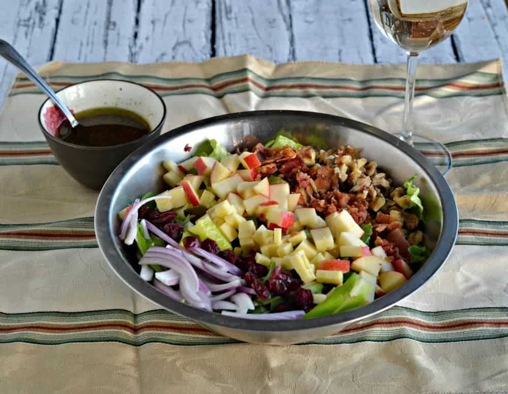 A salad sitting in a stainless bowl, filled with diced apples, diced cheddar, nuts, greens, dried cranberries and sliced onions. The bowl is sitting on a yellow napkin with colorful stripes, a glass of white wine in the background and extra dressing in a dish next to the salad.