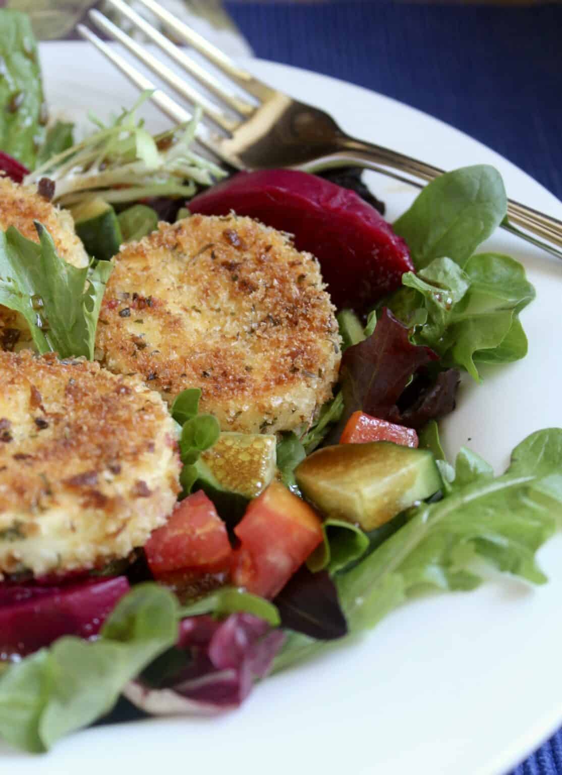 An up close photo of salad greens topped with sliced beets, diced cucumbers, tomatoes, and topped with three breaded rounds of goat cheese. The salad is served on a white plate with a fork to the side.