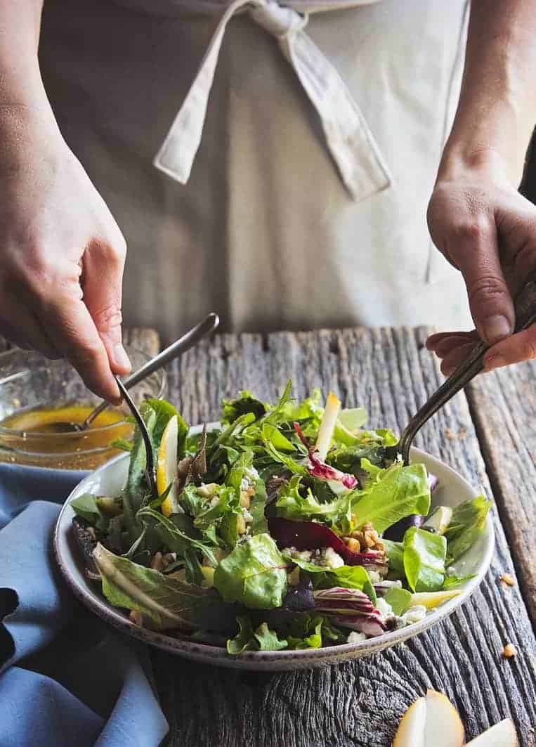 A person wearing an apron holding two utensils, tossing the salad on a wooden table. Salad is in a white bowl with greens, red lettuce, pears, chunks of cheese, and chopped nuts. Extra dressing if off to the side. 
