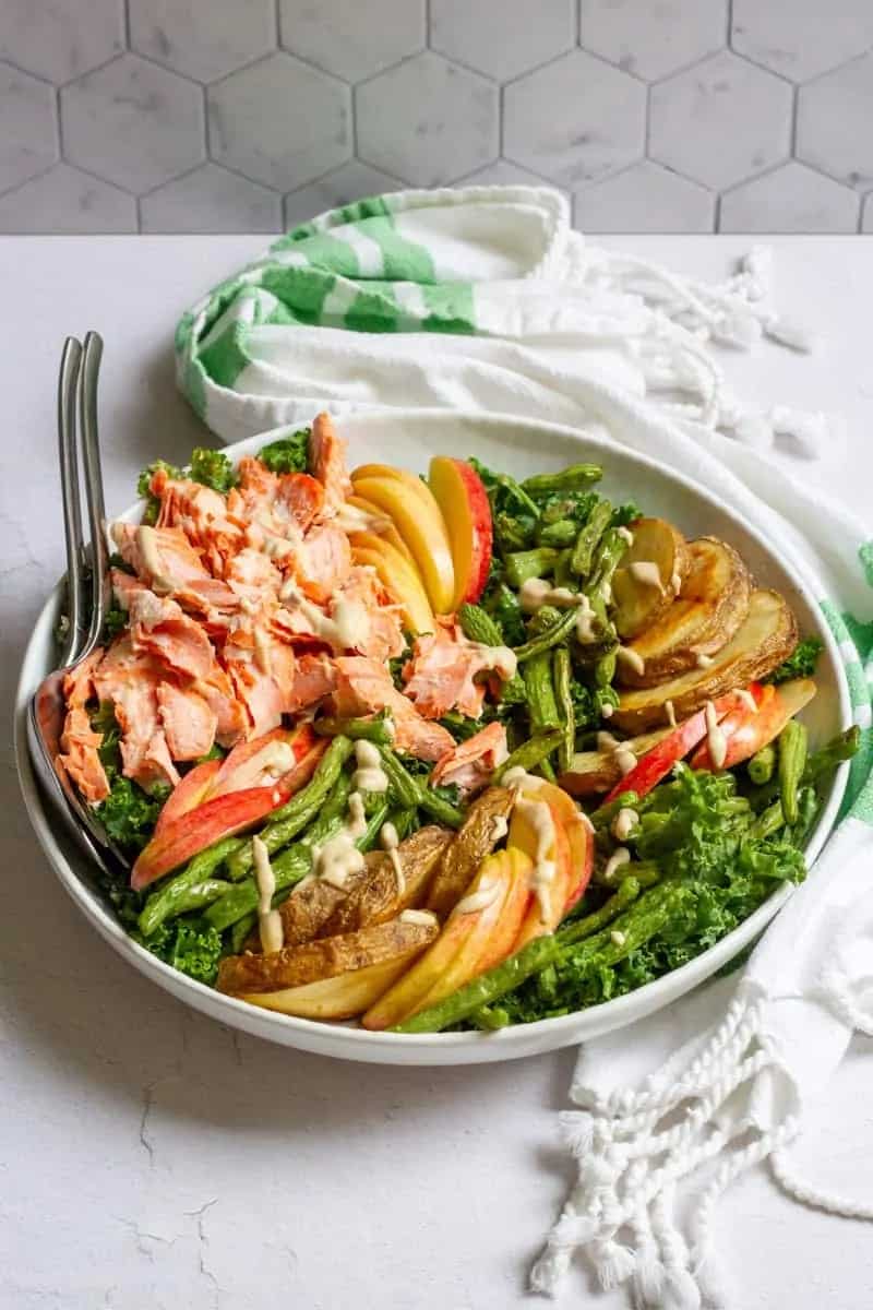 A white bowl sitting on a green striped towel filled with salad ingredients including flaked salmon, sliced potatoes, greens, apples and green beans. The salad is drizzled with a creamy dressing. Serving utensils are sticking out of the bowl. 