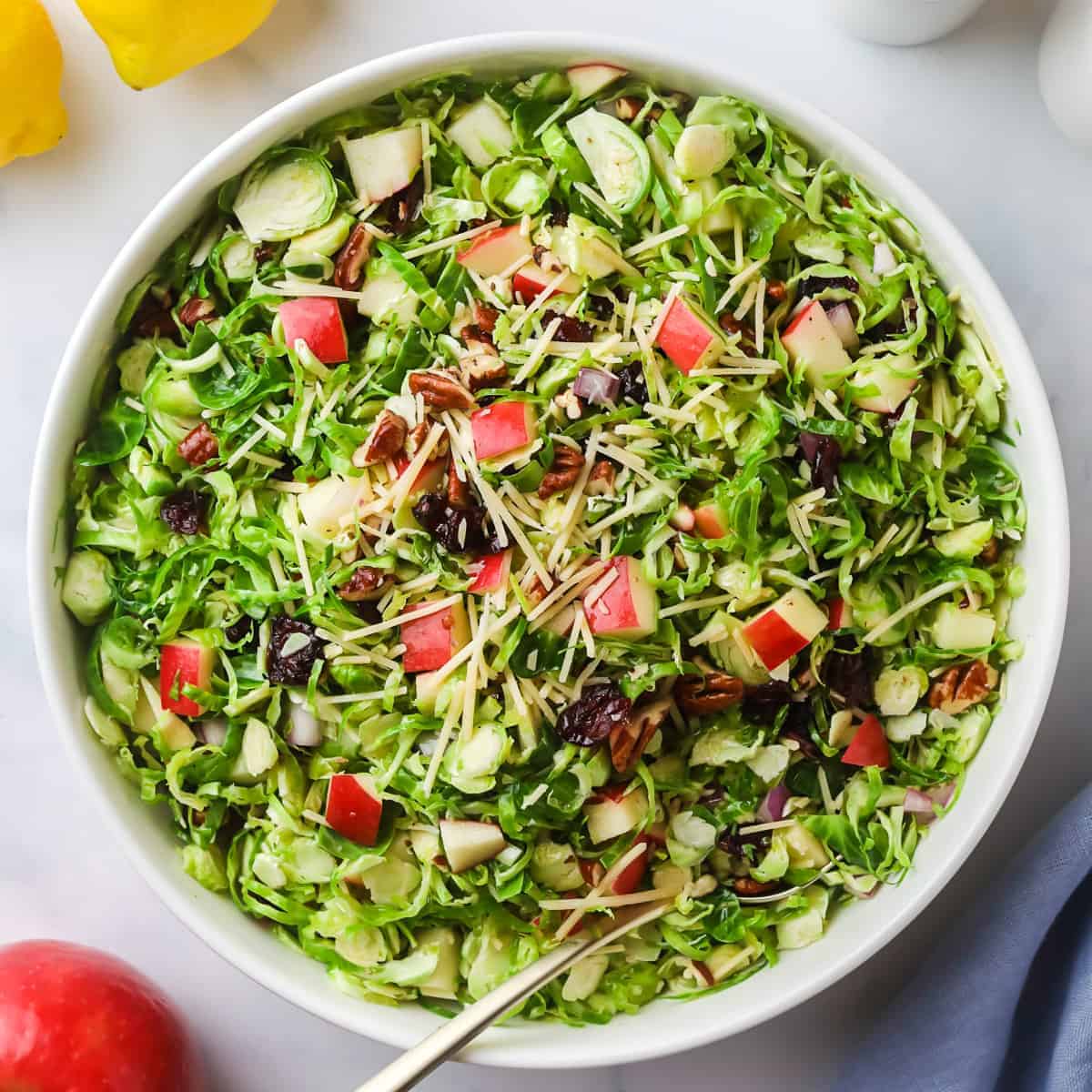 Shaved brussels sprouts in a white bowl topped with pecans, diced red apples, shredded parmesan cheese and dried cranberries. A red apple, lemons and a blue towel surround the bowl. A serving utensil sits inside the salad.