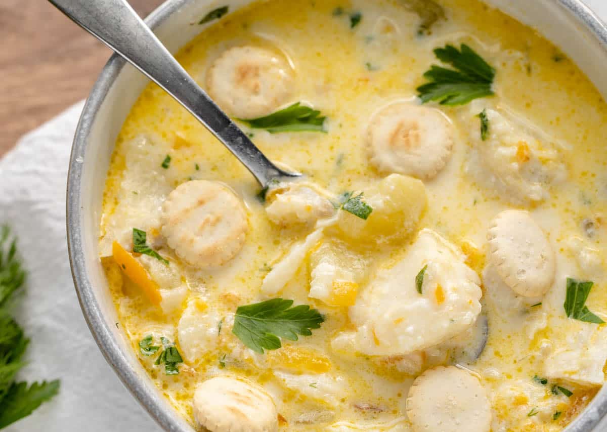 A bowl of fish chowder with chunks of fish, potatoes, herbs, and oyster crackers. A spoon sits in the chowder.