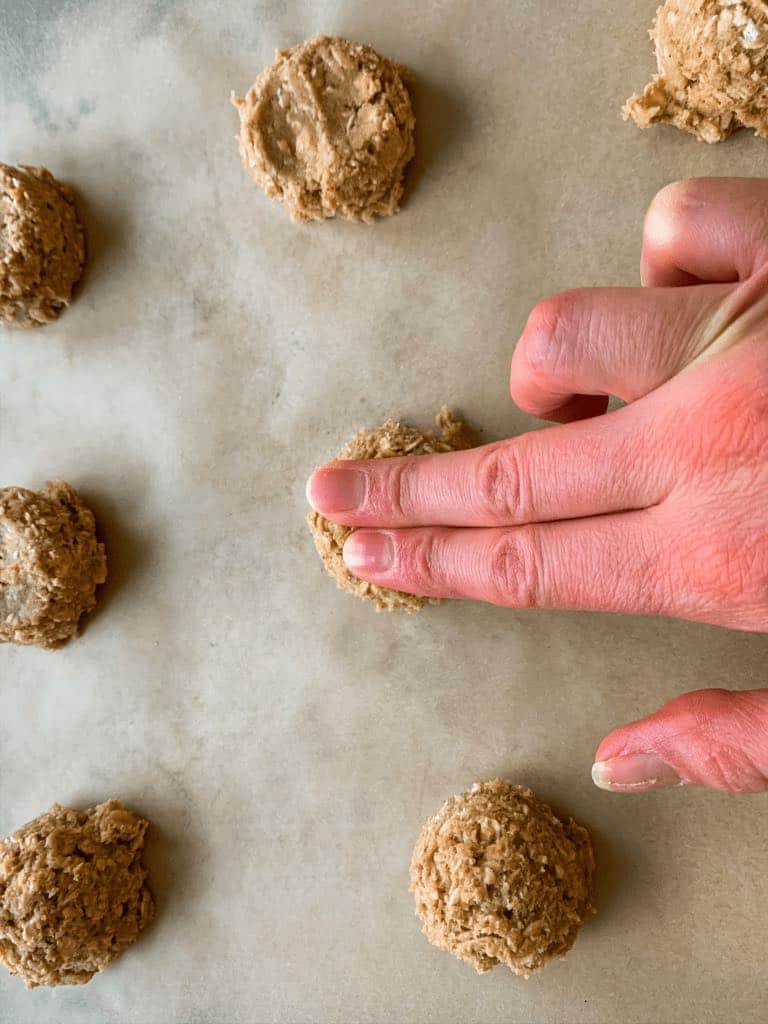 two fingers pressing the center of a cookie dough ball to flatten it slightly. On a parchment paper lined sheet pan.
