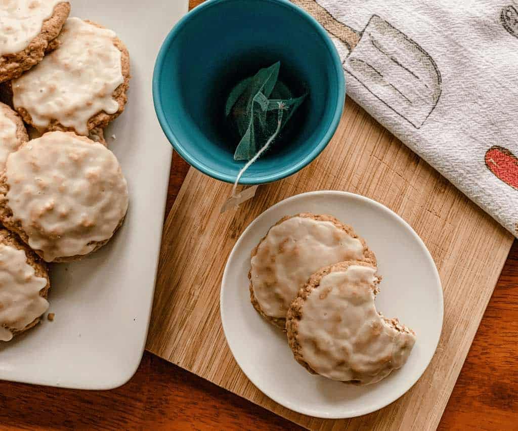 Two iced oatmeal cookies on a small plate, next to a blue mug and a white platter of iced oatmeal cookies.