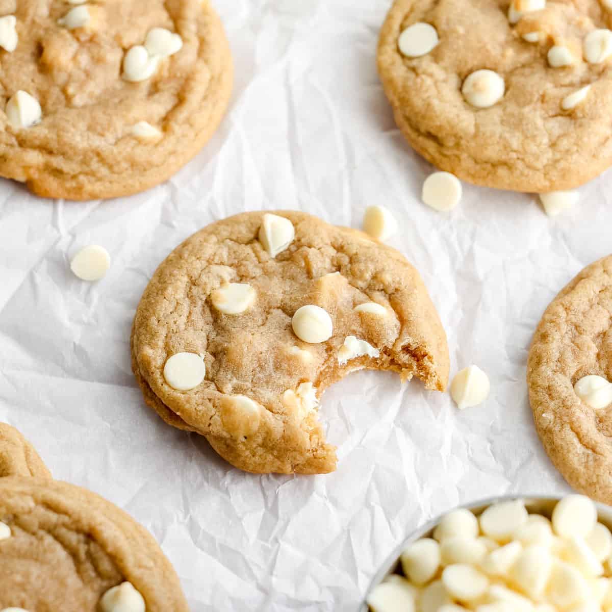 A bitten white chocolate chip cookie on a white surface. Other cookies and white chocolate chips surround it.