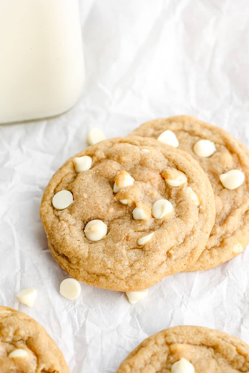 Two white chocolate chip cookies sitting on top of each other. Additional cookies and a glass of milk in the background.