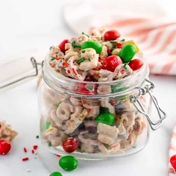 A jar of white chocolate Chex mix with red and green M&Ms and sprinkles on top and around the table. A napkin in the background.