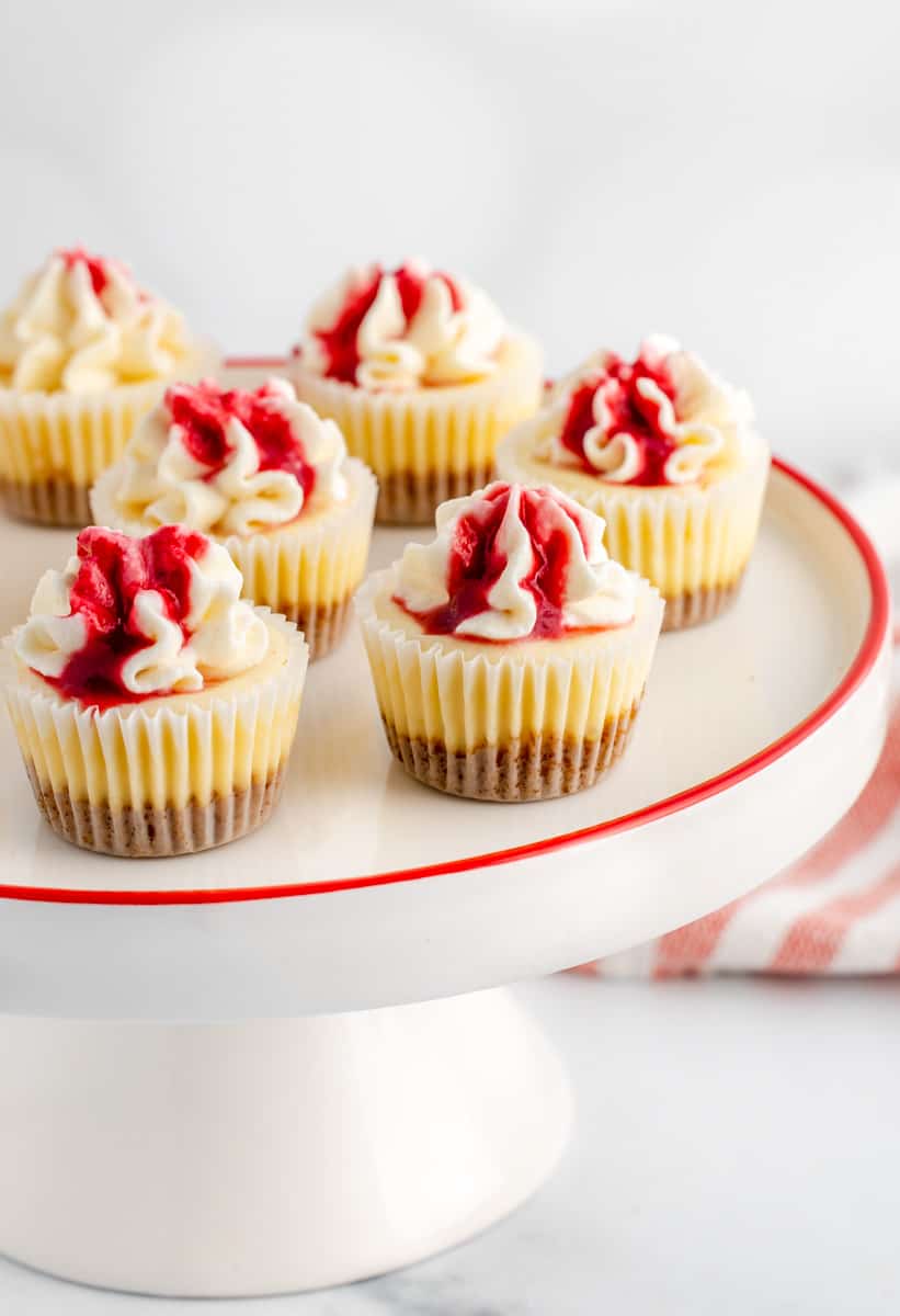 A red and white cake stand with mini cheesecake bites, topped with red fruit juice and whipped cream. A red stripe napkin in the back.