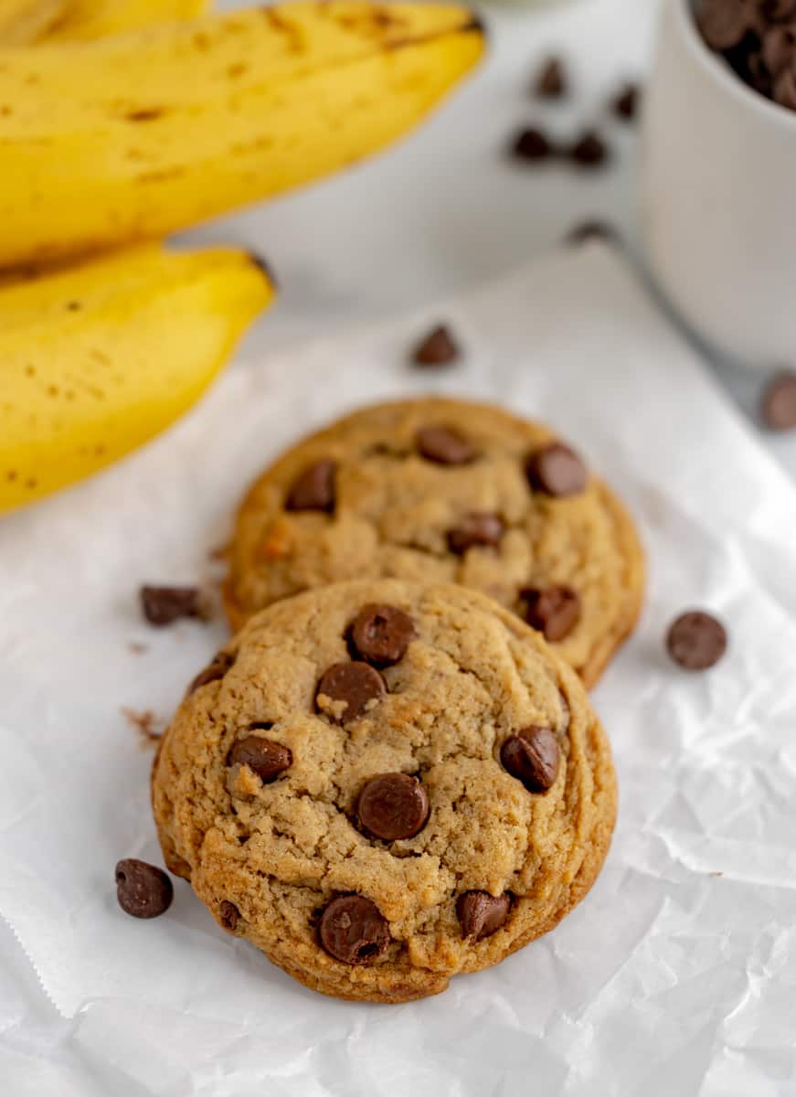 Two chocolate chip cookies with bananas and chocolate chips in the background.