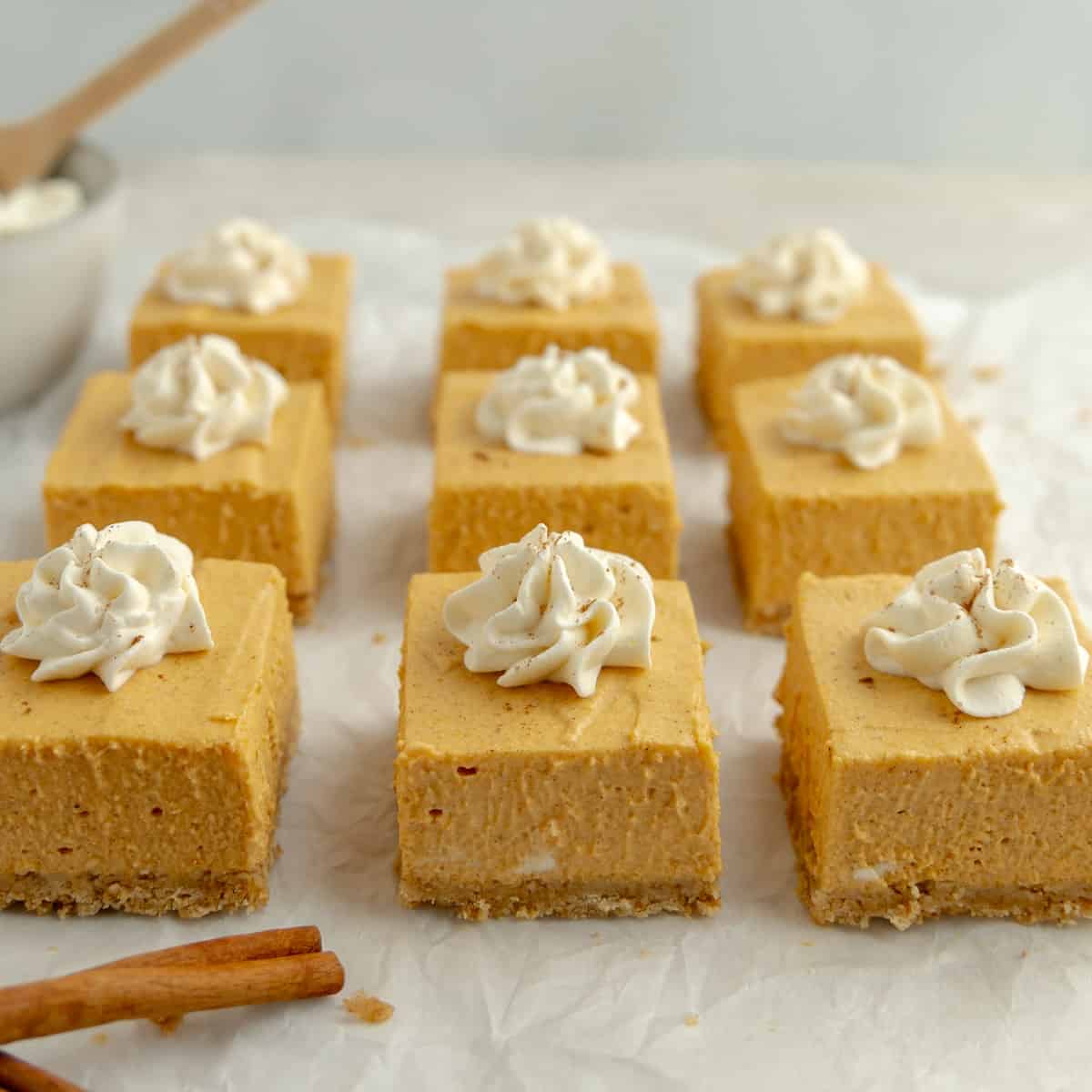 A group of pumpkin cheesecake squares topped with whipped cream. Cinnamon sticks on the side.