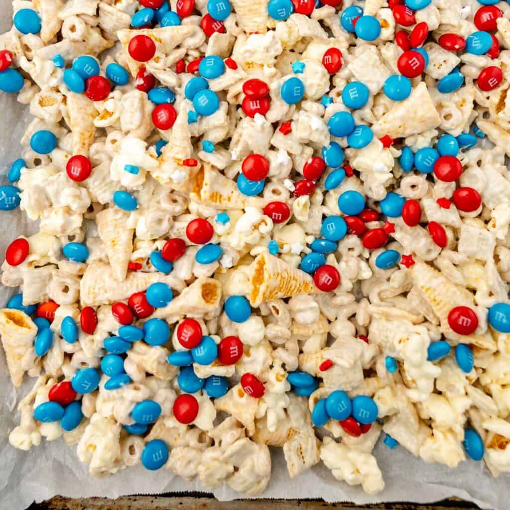 A close-up of a snack mix with popcorn, cereal, and red and blue M&Ms on a sheet of parchment paper.