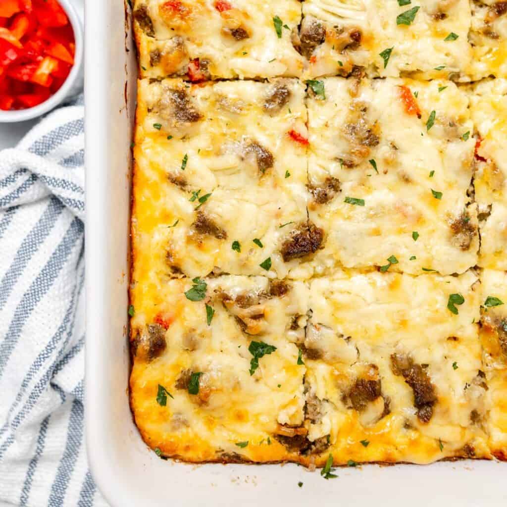A baked breakfast casserole with sausage, cheese, and vegetables cut into squares, in a white dish on a striped towel, with a small bowl of diced red bell peppers nearby.