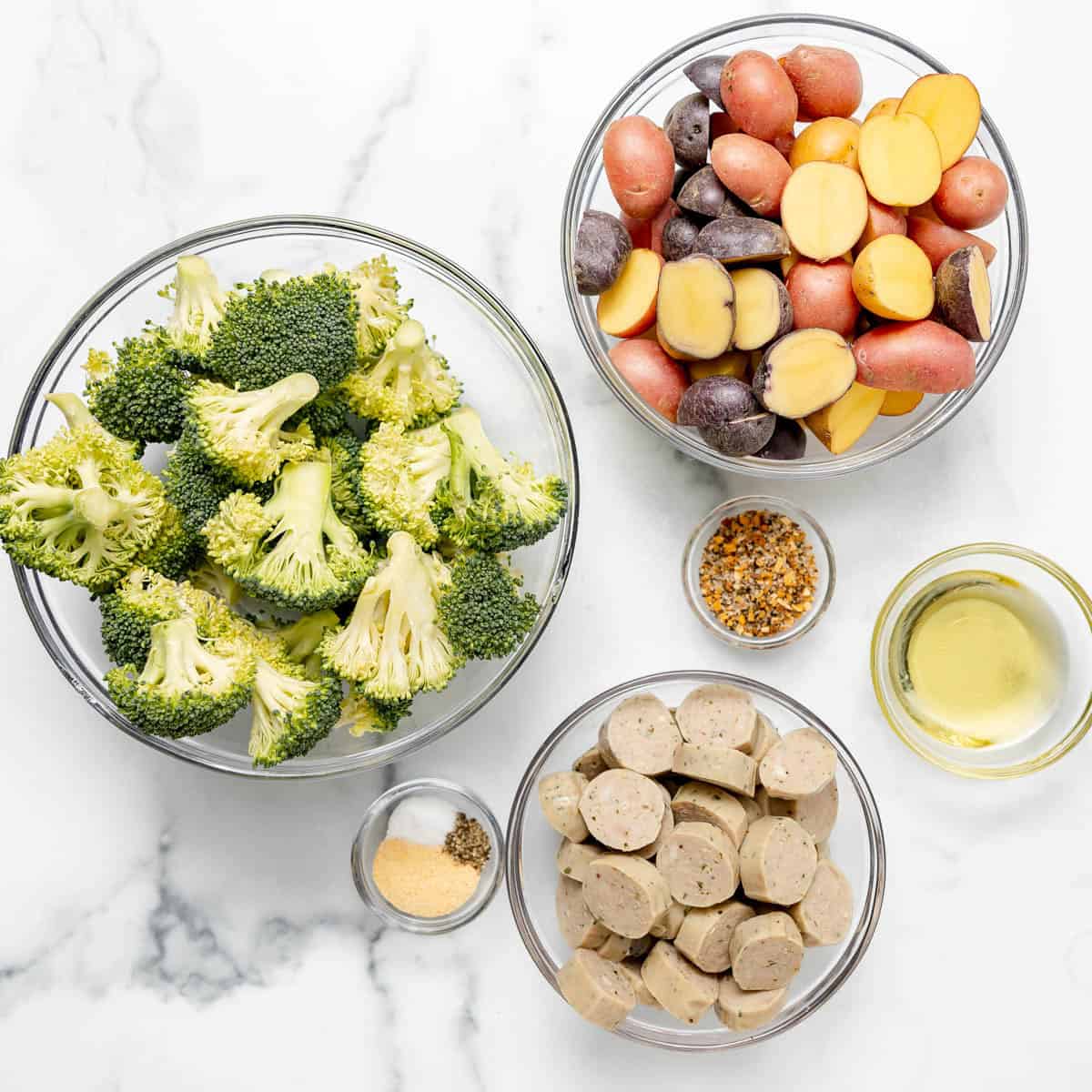 Five clear bowls on a marble surface containing broccoli florets, halved baby potatoes, sliced sausage, spices, and a small amount of oil.
