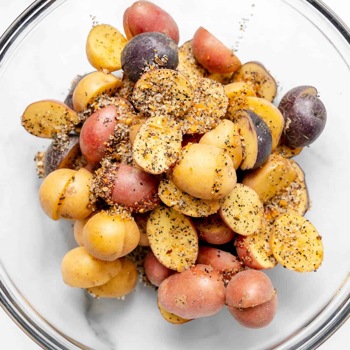 Halved red, yellow, and purple baby potatoes tossed with seasoning and coarse salt in a glass bowl.