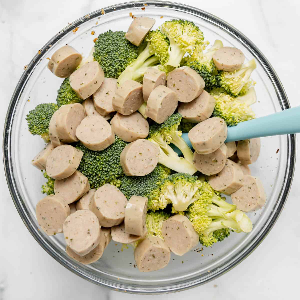 Sliced sausage and broccoli florets in a glass mixing bowl with a blue spatula on a white surface.
