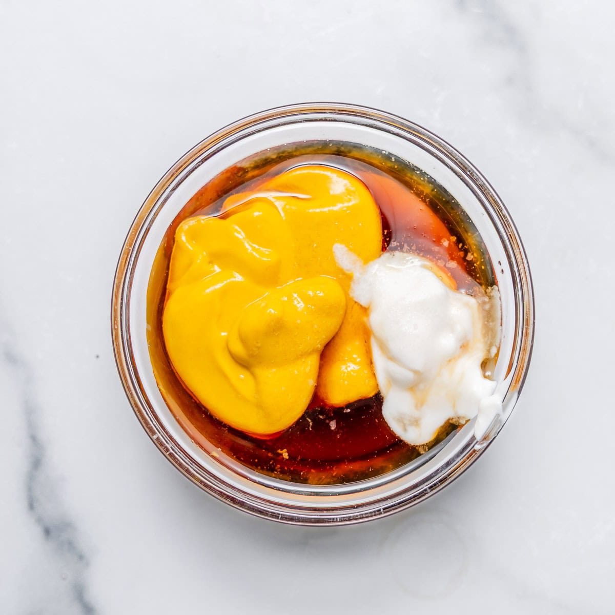 A glass bowl containing yellow mustard, yogurt, and maple syrup, on a white marble surface.