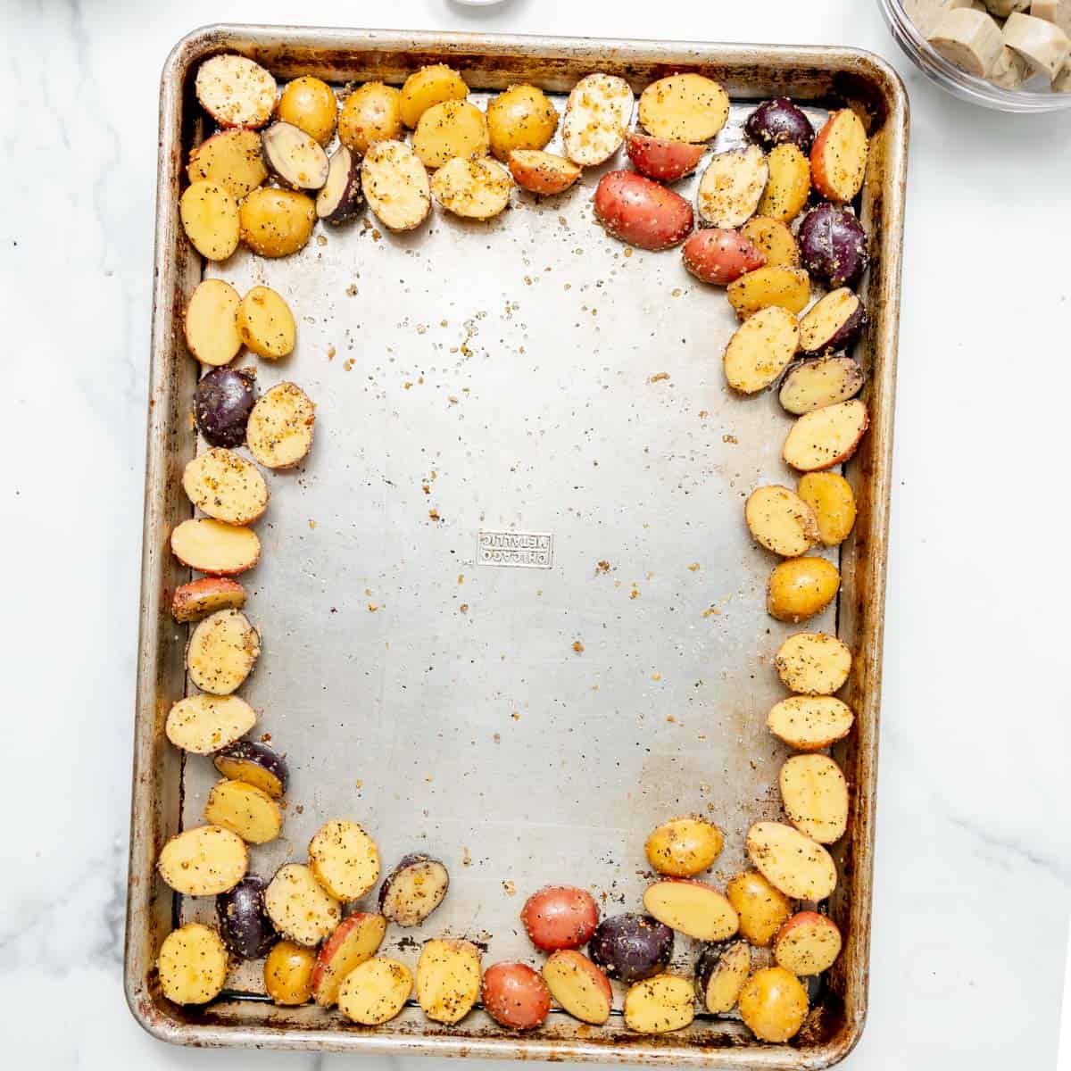 A baking sheet with halved, seasoned baby potatoes arranged around the edges, leaving the center empty, on a white surface.