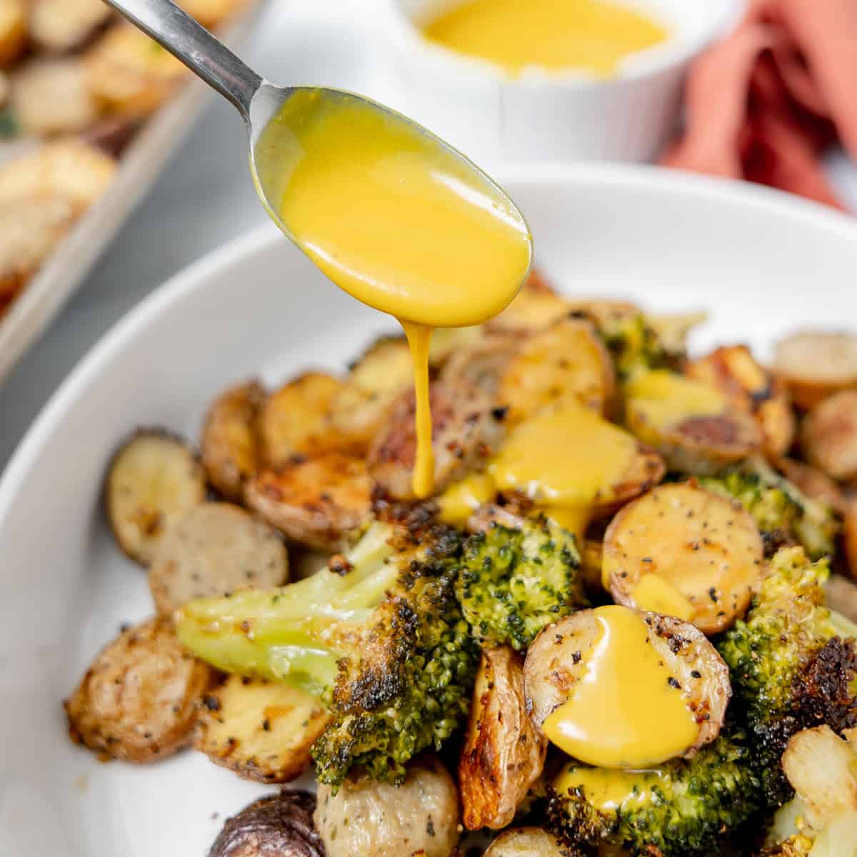 A spoon drizzles yellow mustard sauce over roasted potatoes and broccoli on a white plate, with a ramekin of sauce in the background.