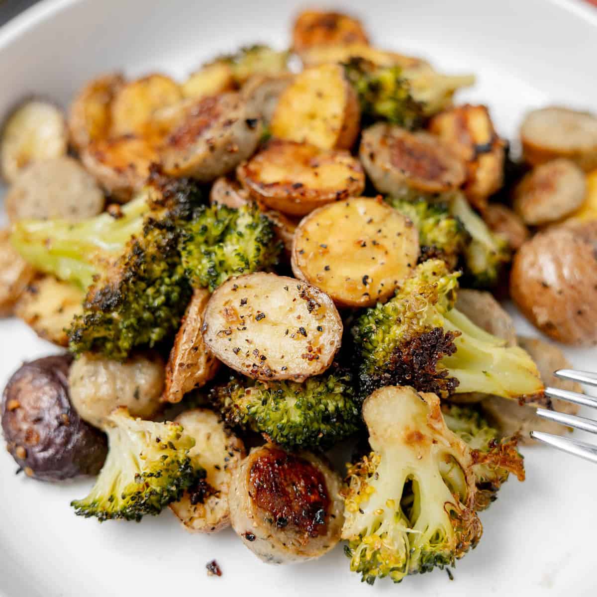 A white bowl filled with roasted broccoli florets and sliced baby potatoes, seasoned with herbs and pepper. A fork rests at the side of the bowl.