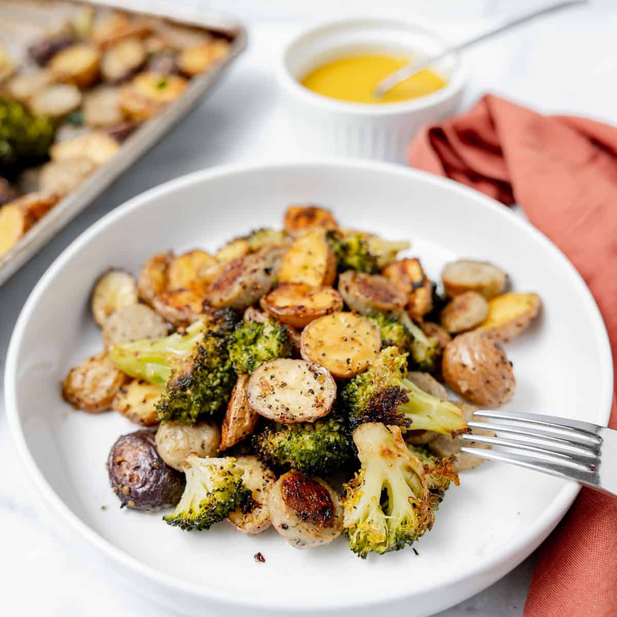 A white plate with roasted broccoli and baby potatoes, a fork resting on the side, and a small dish of yellow sauce with a spoon in the background.