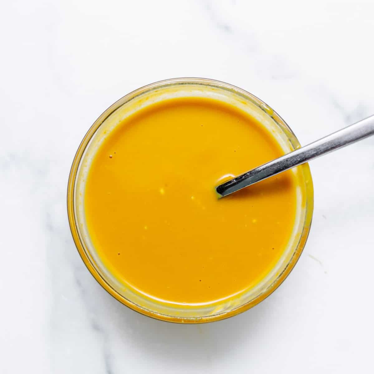 A small glass bowl filled with yellow-orange sauce, with a metal spoon resting inside, on a white marble surface.