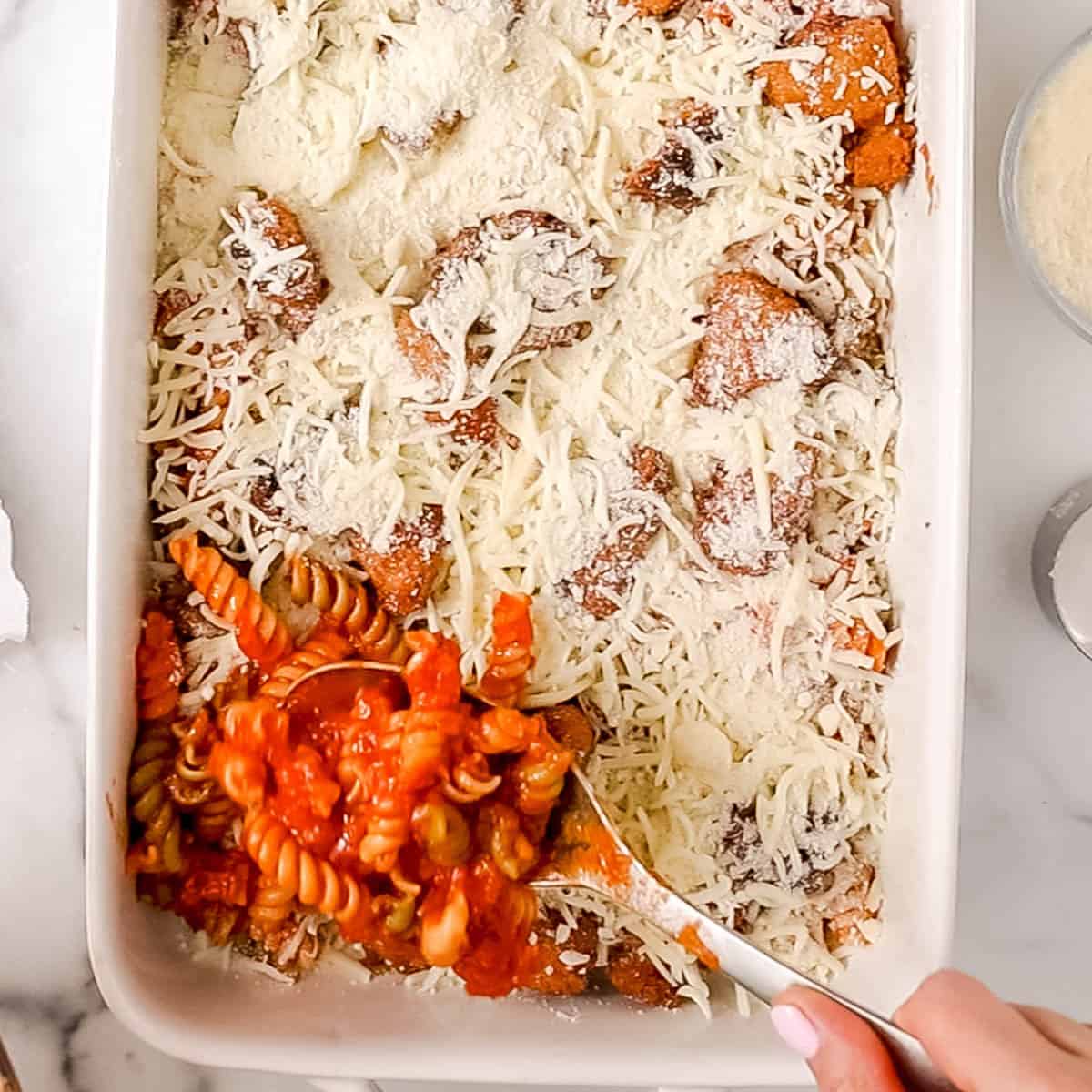 Hand spooning rotini pasta with tomato sauce into a white baking dish, topped with shredded cheese and meat.