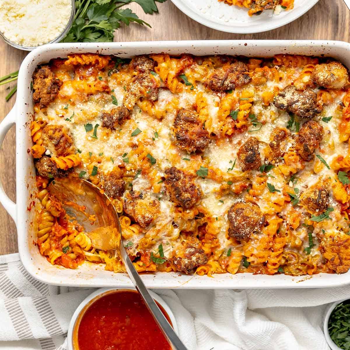 A baked pasta casserole with chicken, melted cheese, and herbs in a white dish, with a serving spoon and bowls of marinara sauce and grated cheese nearby.
