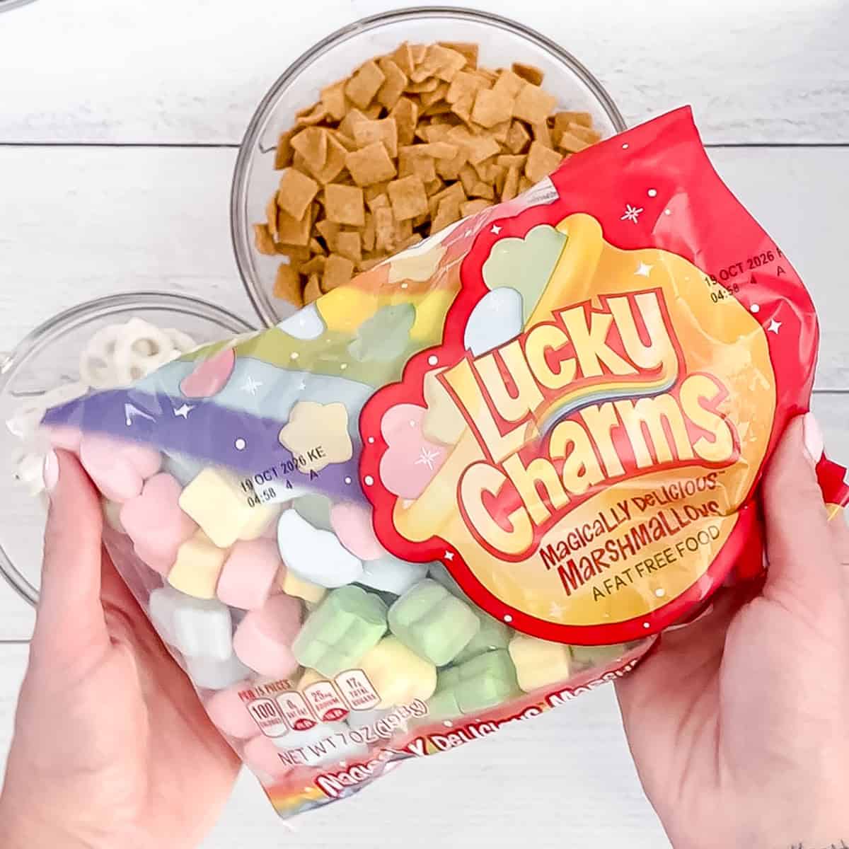 A person holds a bag of Lucky Charms marshmallows over bowls filled with cereal pieces and white candy melts on a white surface.