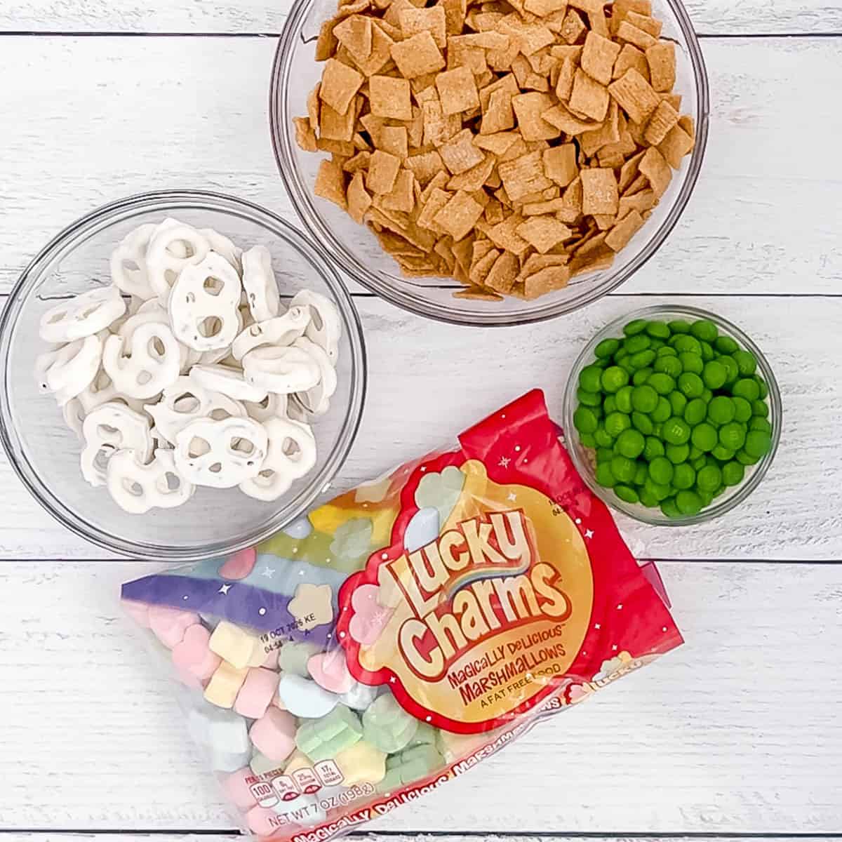 Bowls of cereal squares, yogurt-covered pretzels, green candy-coated chocolates, and a bag of Lucky Charms marshmallows on a white wooden surface.