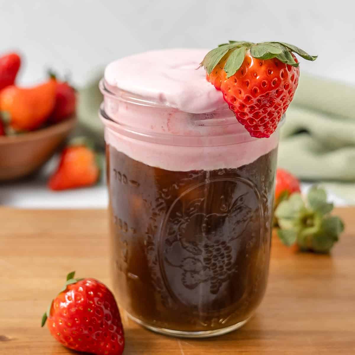 A glass jar filled with iced coffee topped with pink foam and garnished with a fresh strawberry, with more strawberries in the background.